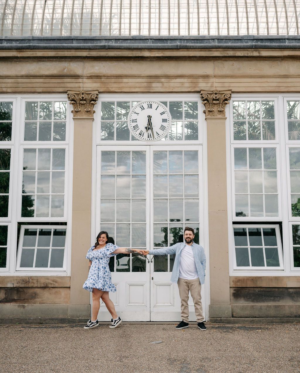 Steph and Jonny holding hands in front of a glasshouse with a large clock above the doors